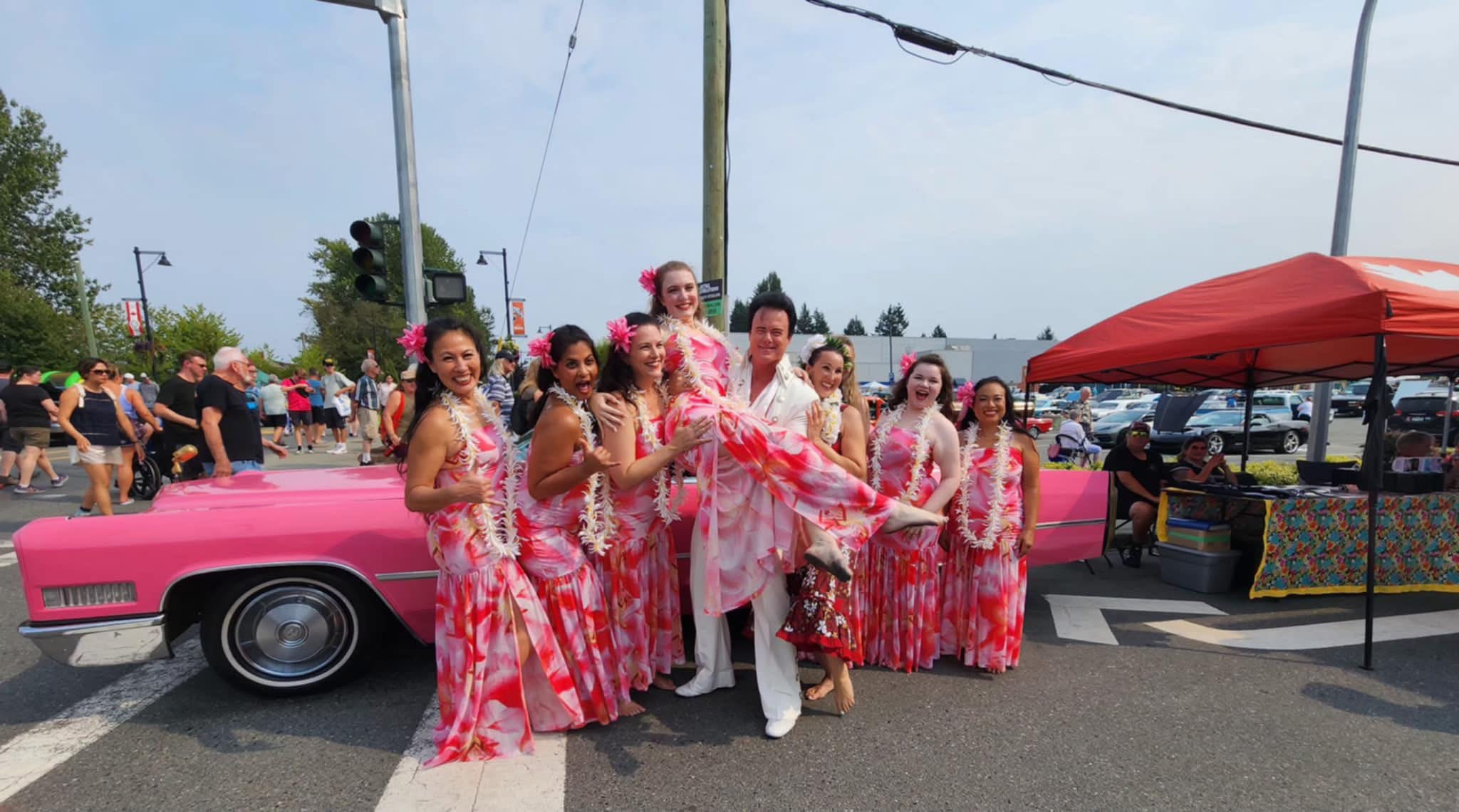 Gina (second from left) with her hula troupe in pink and white floral dresses, posing next to a pink Cadillac at a summer event.