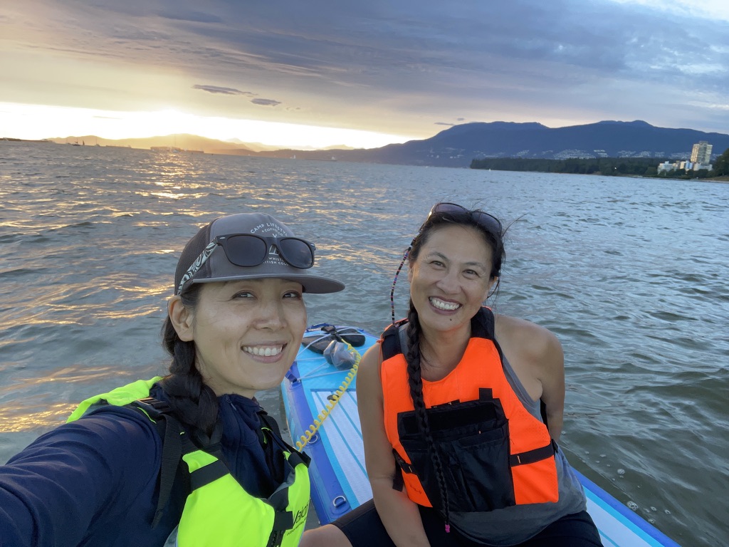 Gina and a friend on a paddleboard in life vests, laughing, with the sun setting over the ocean behind them.