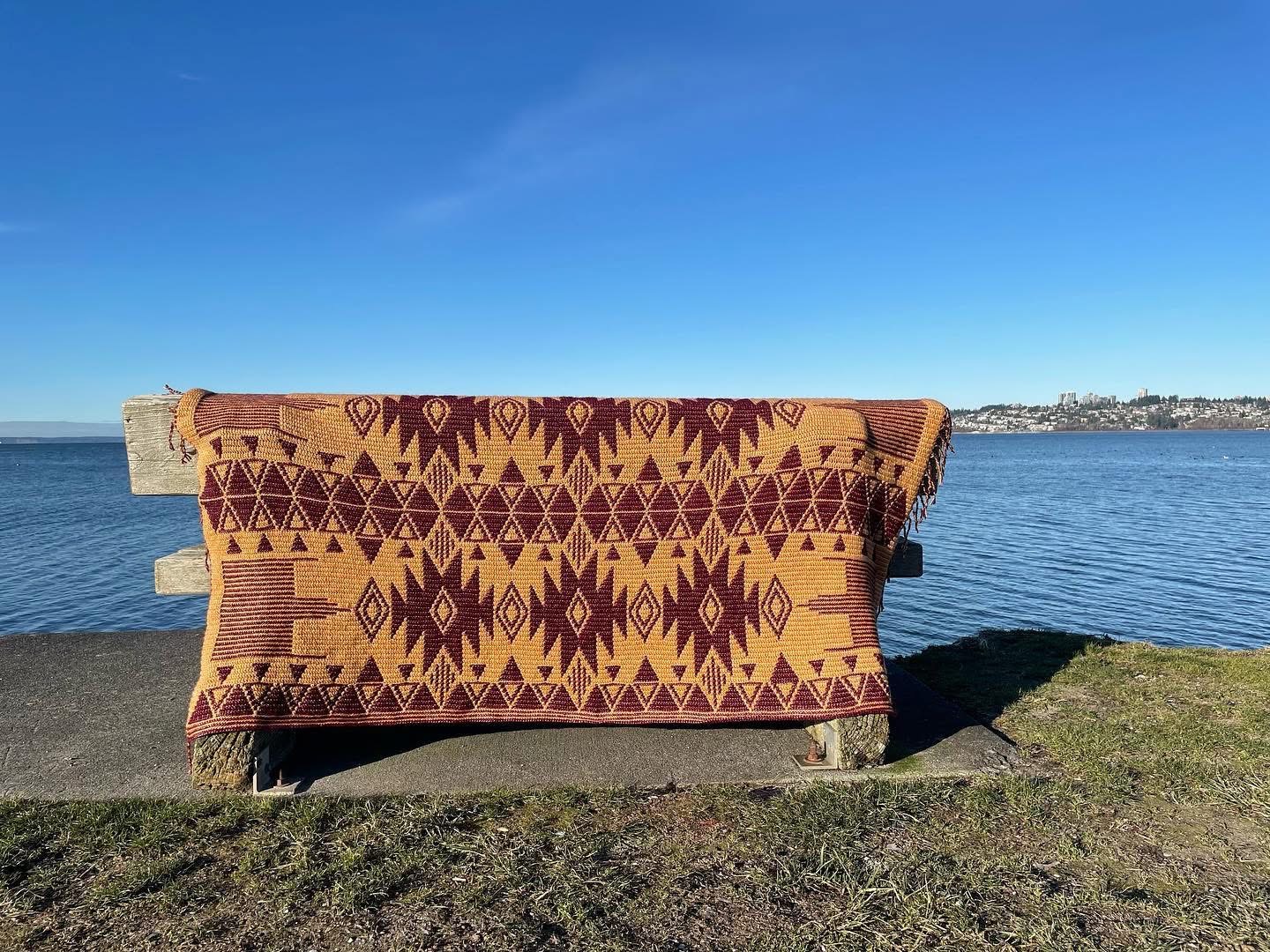 A burgundy and ochre geometric crochet blanket draped over a driftwood bench by the sea, blue sky and skyline behind.